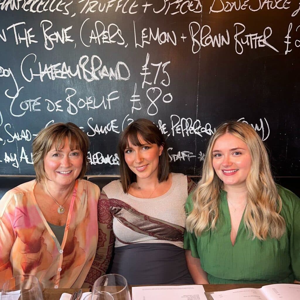 Three women smiling at a restaurant with a chalkboard menu in the background.