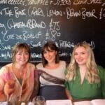 Three women smiling at a restaurant with a chalkboard menu in the background.