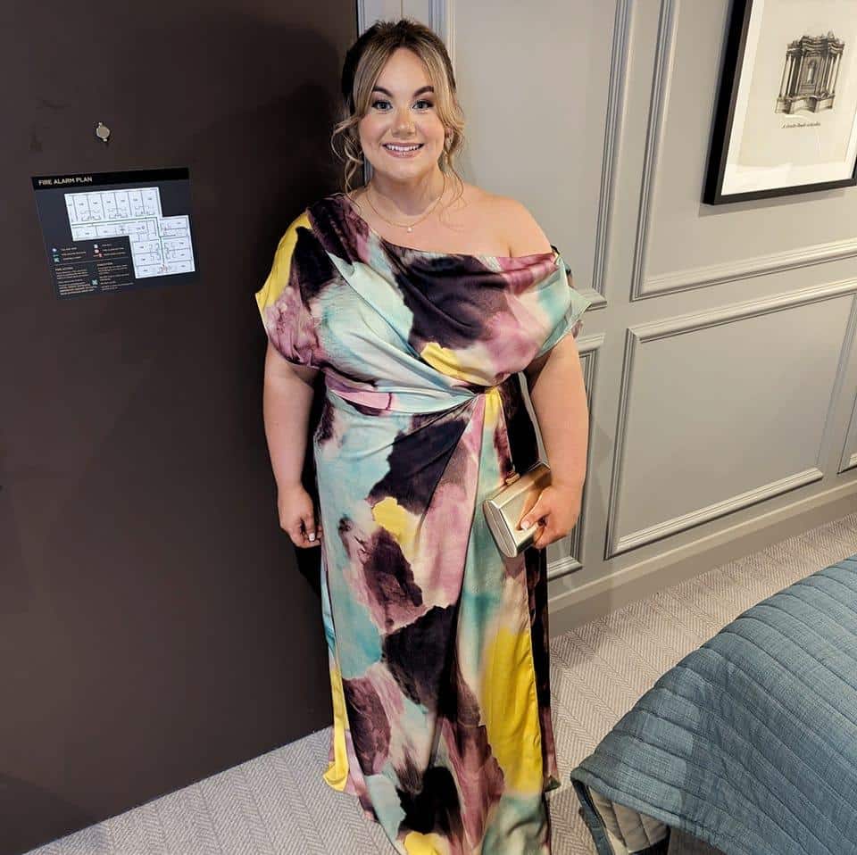 Smiling woman in a colorful off-shoulder dress holding a clutch in a hotel room.