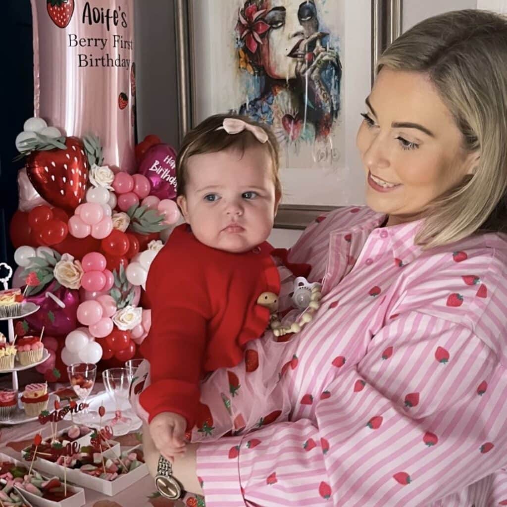 Woman holding baby at birthday party with pink and red decorations and balloons.