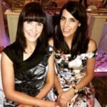 Two women seated, smiling, one in a black patterned dress and the other in a floral off-shoulder dress, indoors.