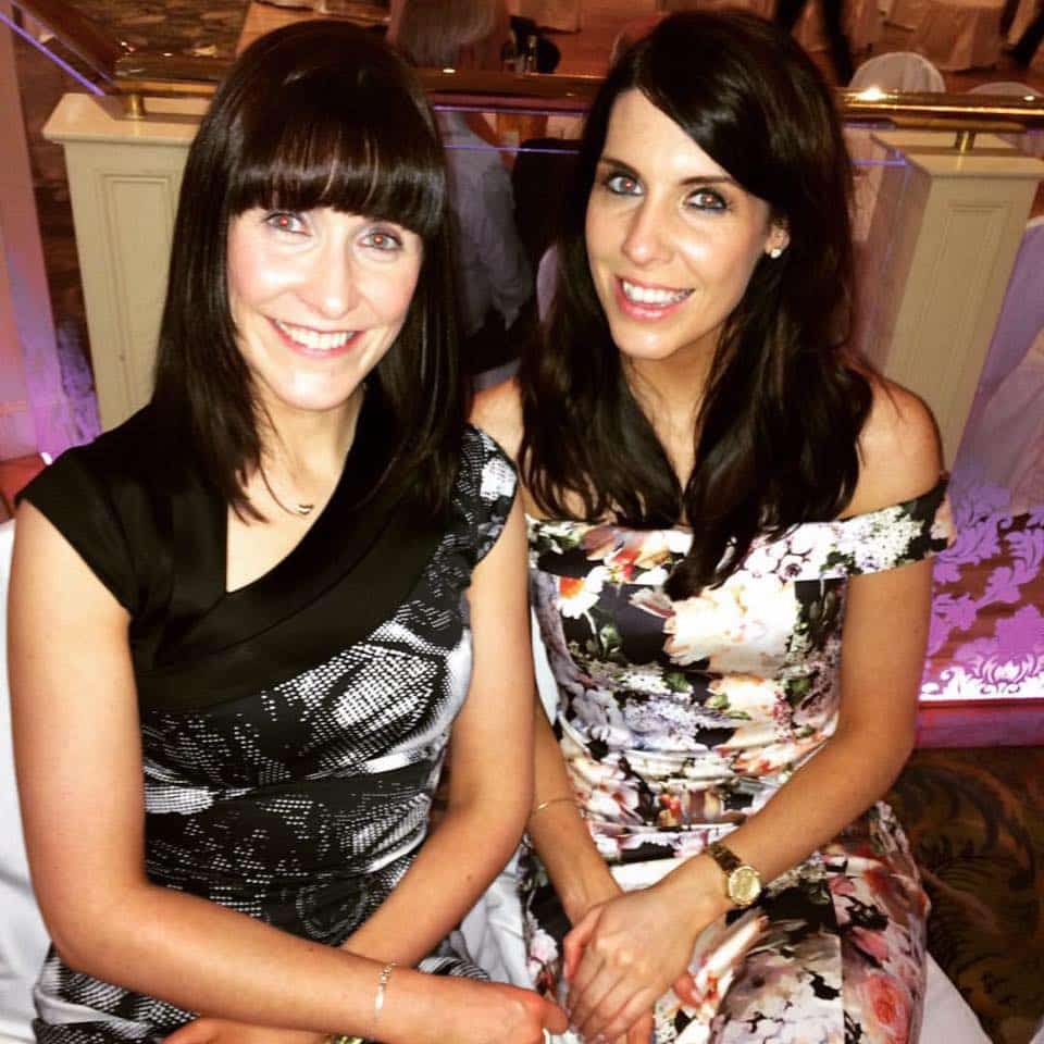 Two women seated, smiling, one in a black patterned dress and the other in a floral off-shoulder dress, indoors.