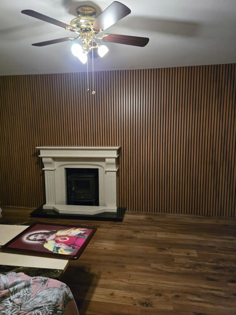 Room with wooden paneling, ceiling fan, white fireplace, and a table with religious artwork.