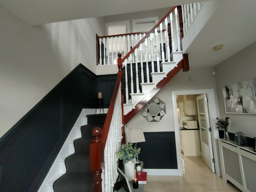 Modern interior with a staircase, white walls, dark paneling, decorative mirror, and a glimpse of a kitchen.