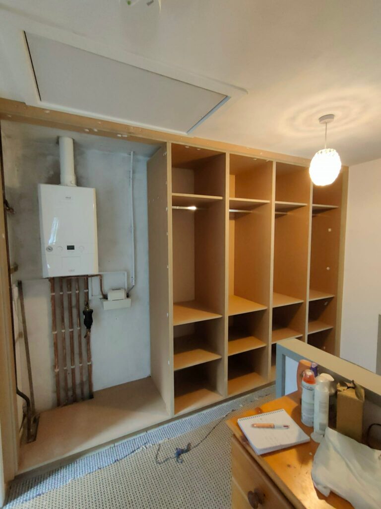 Unfinished closet with open shelving, exposed boiler, and a ceiling light in a room under renovation.