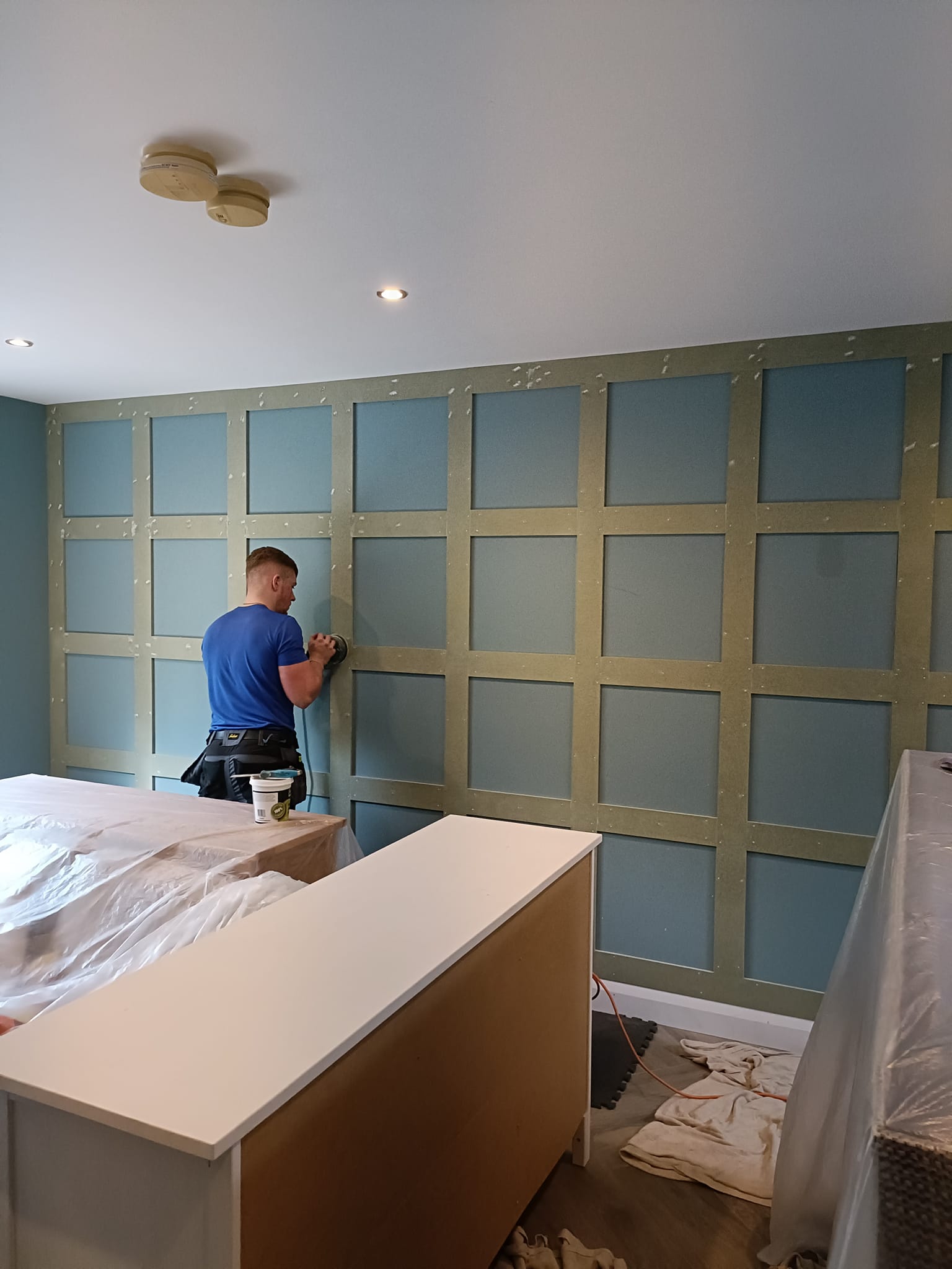 Person working on a textured wall in a room, surrounded by covered furniture and construction materials.
