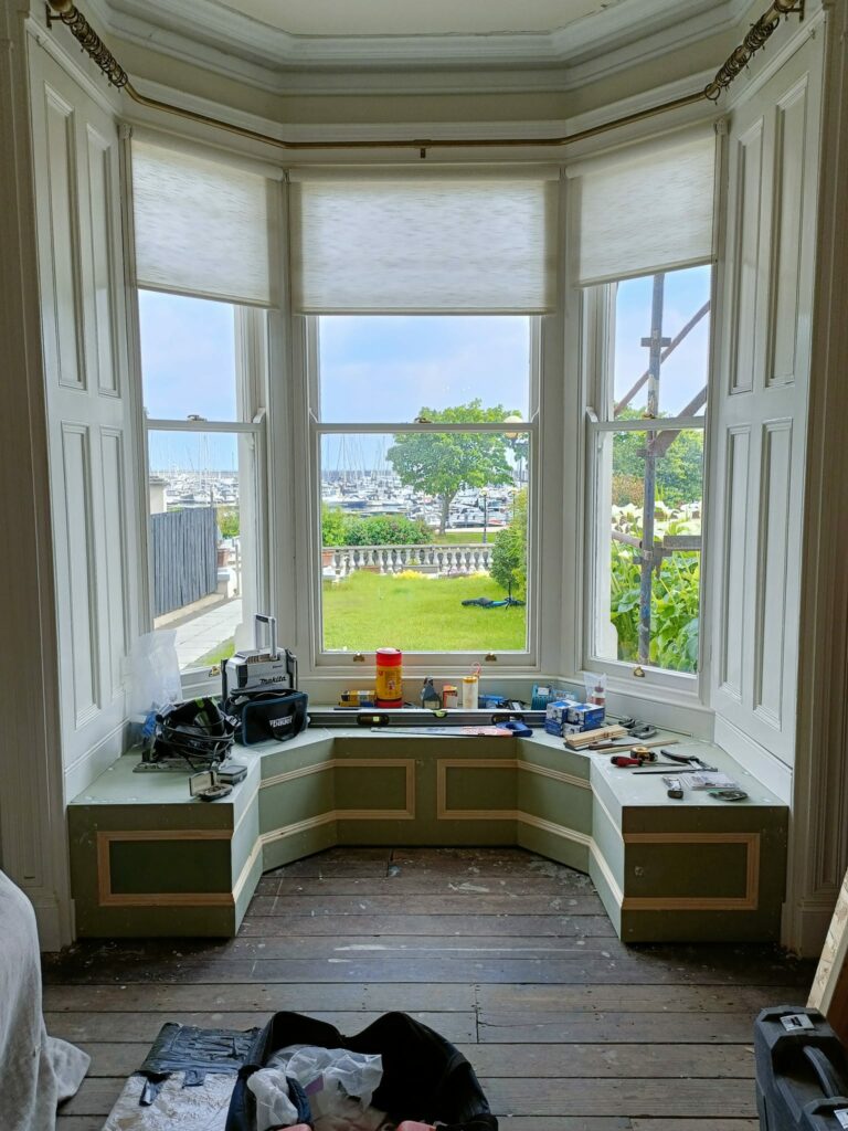Bay window with view of garden and harbor, surrounded by tools and wooden benches indoors.