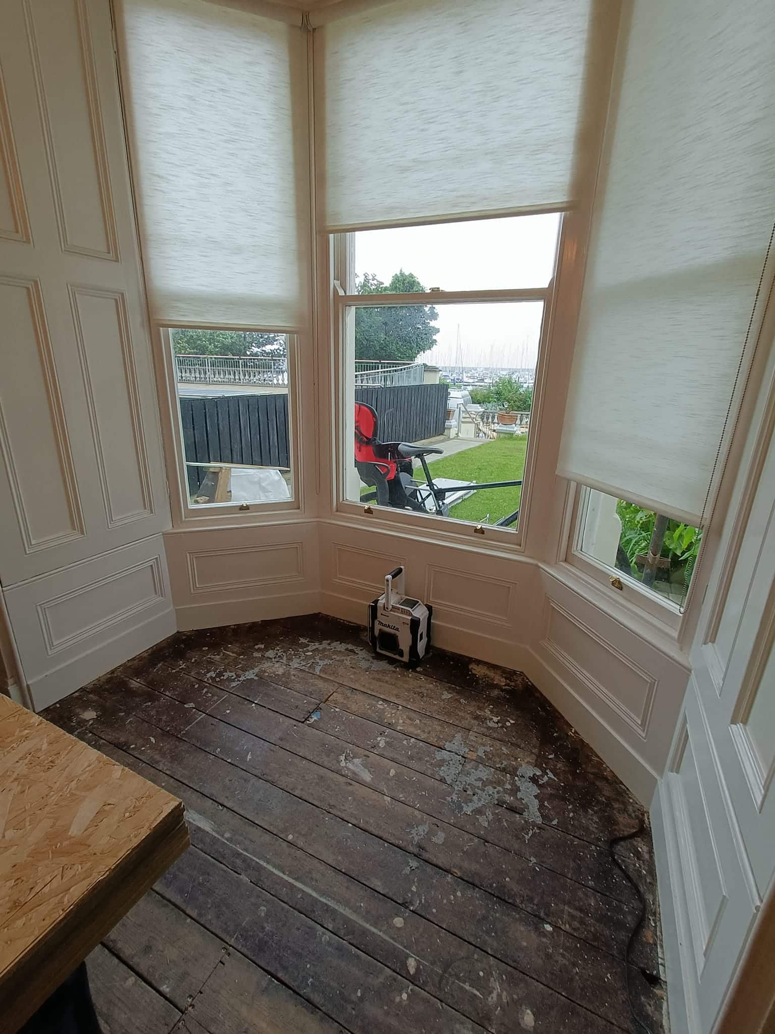 Bay window with roller blinds, overlooking garden and water, old wooden floor, and toolbox on the floor.