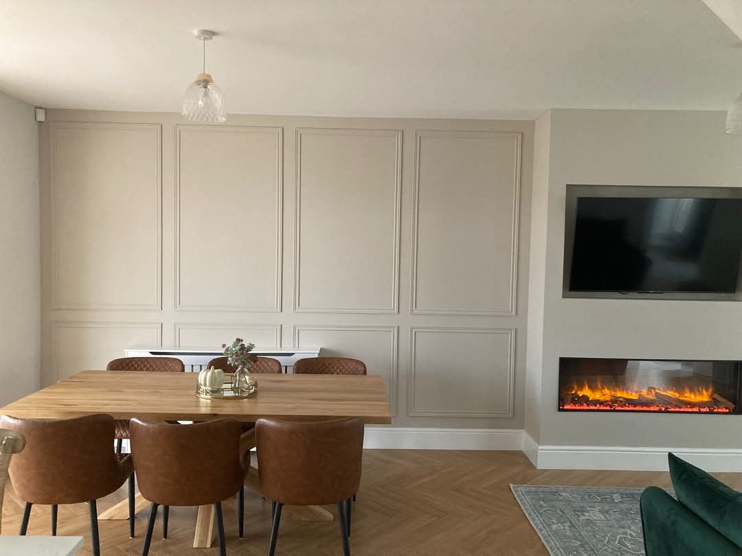 Dining room with beige paneled walls, wooden table, brown chairs, and a modern fireplace beneath a wall-mounted TV.