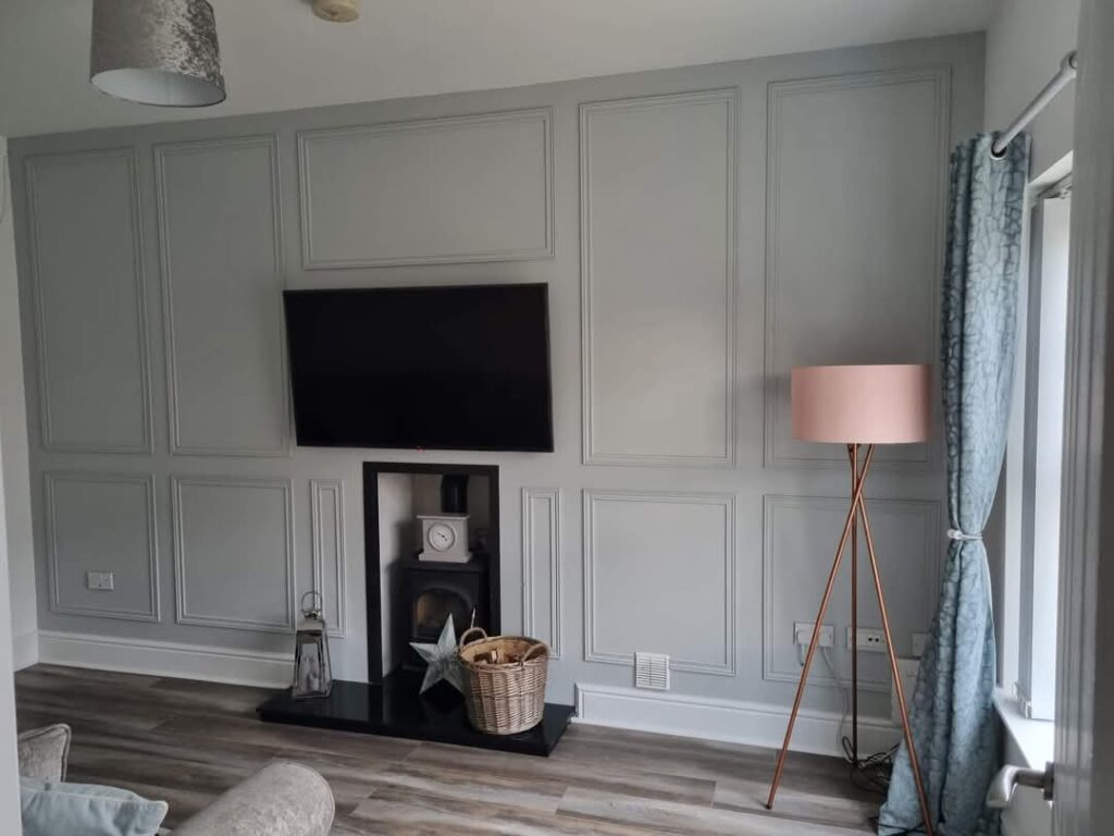 Elegant living room with paneled gray walls, wall-mounted TV, modern lamp, and fireplace, reflecting French-style decor.