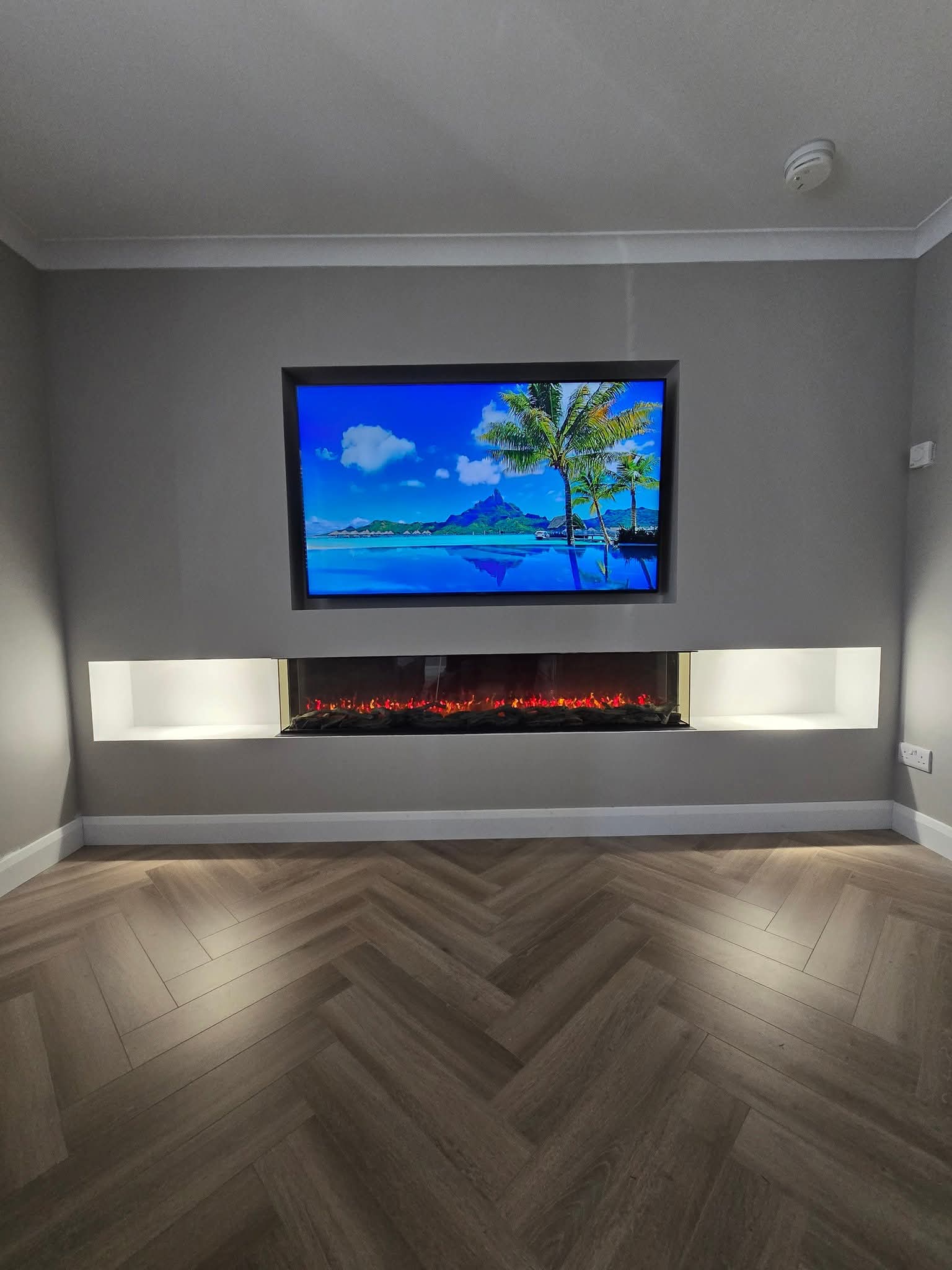 Modern media wall with a widescreen TV above an electric fireplace, set in a minimalist room with herringbone-patterned flooring.