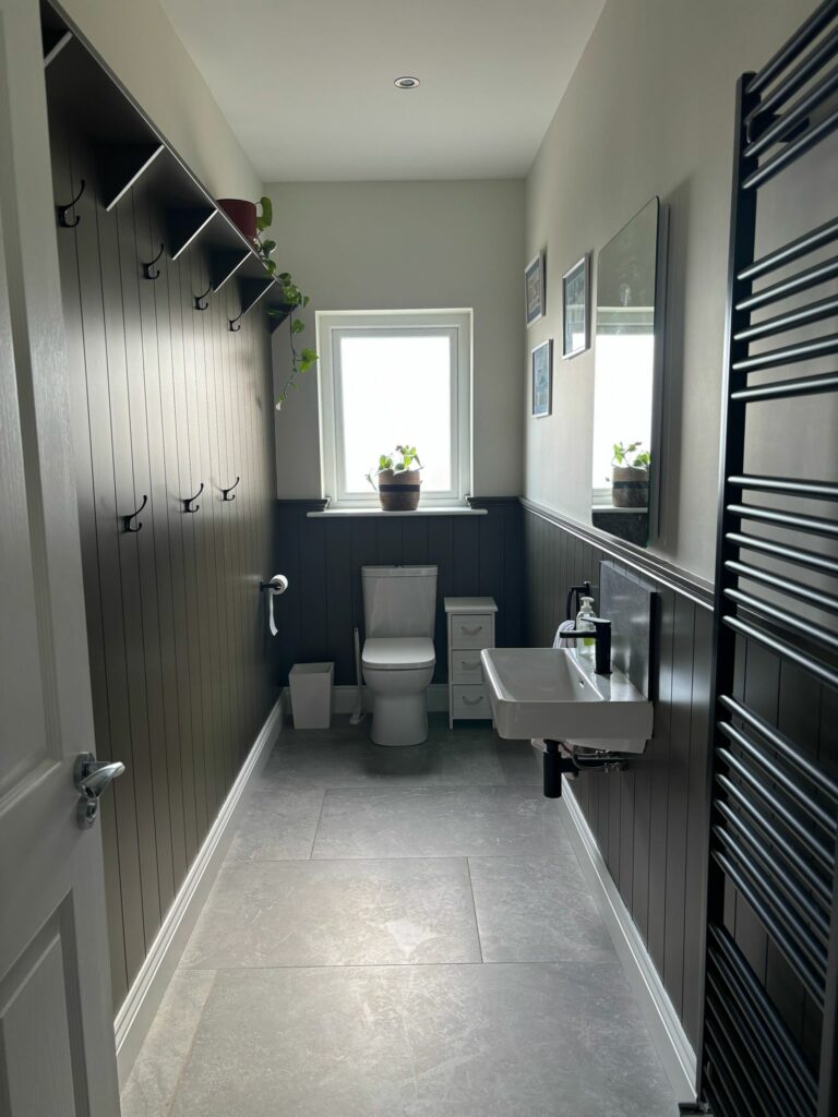 Narrow bathroom with tongue and groove wall paneling, featuring a toilet, sink, window with plant, and wall hooks.