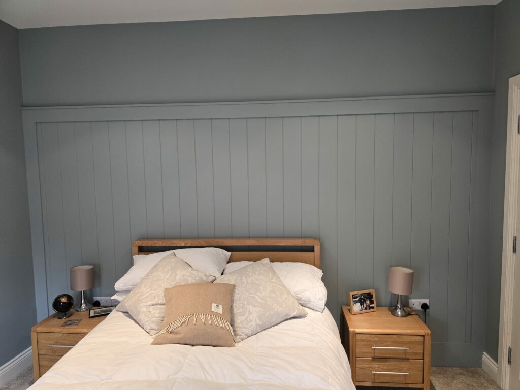 Bedroom with tongue and groove paneling behind a wooden bed, flanked by nightstands and lamps.