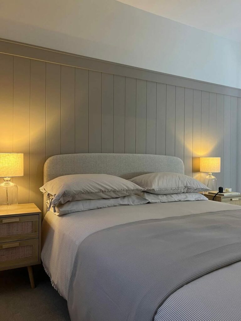 Bedroom with tongue and groove paneling, bed with gray linens, and warm-lit bedside lamps.