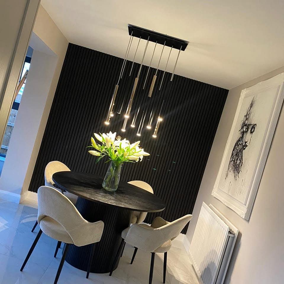 Modern dining room with round table, beige chairs, hanging lights, and a vase of white lilies against a dark accent wall.