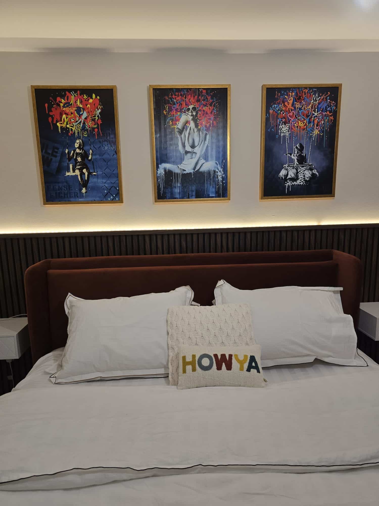 Bedroom with a white bed, decorative "HOWYA" pillow, and three colorful artworks on the wall above.