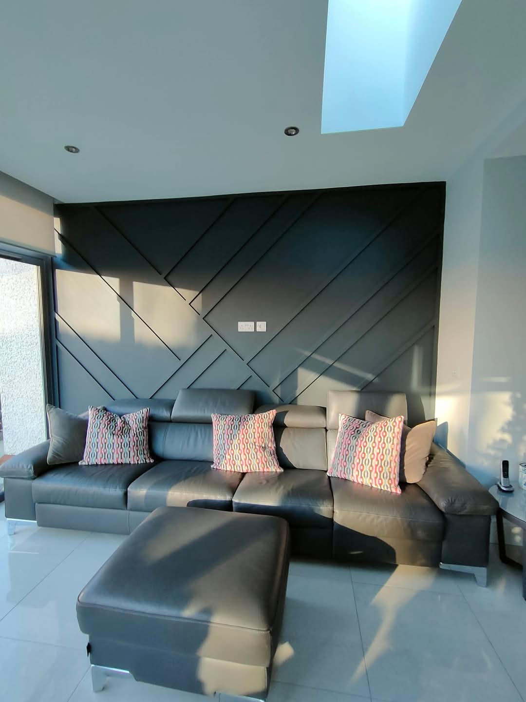 Modern living room with a dark sectional sofa, patterned cushions, and geometric wall paneling, under skylight.