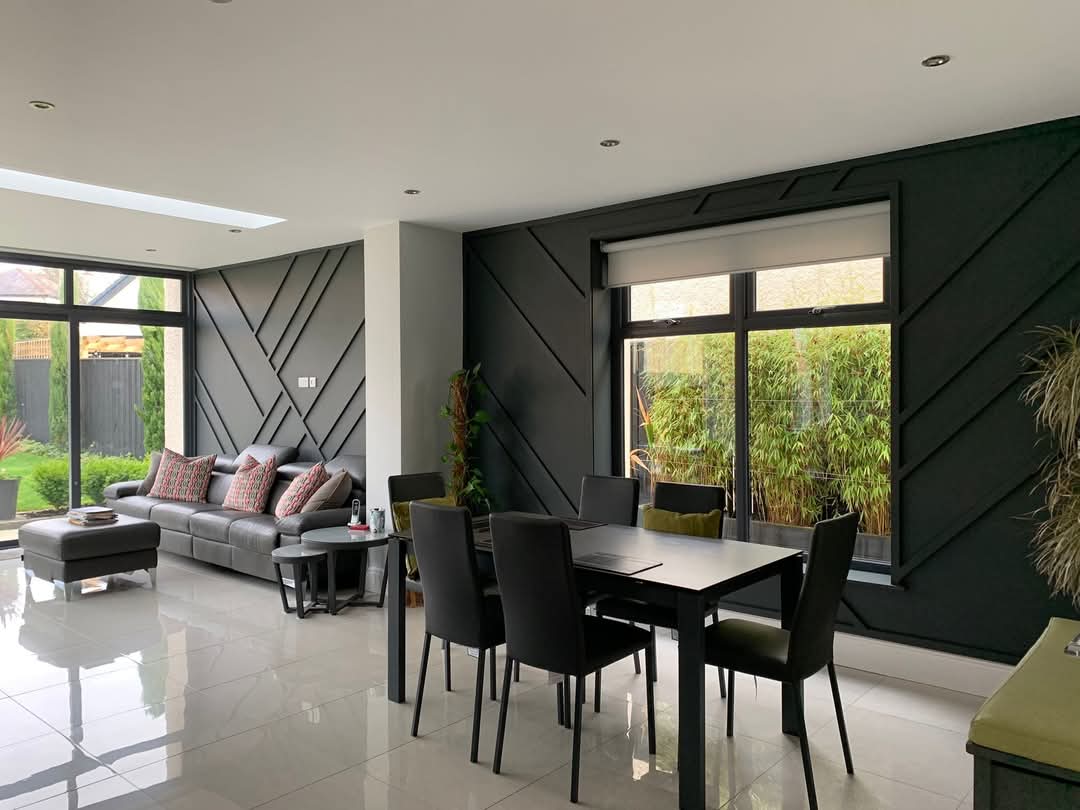 Modern living and dining area with gray sofa, black dining table, geometric wall design, and large windows overlooking greenery.
