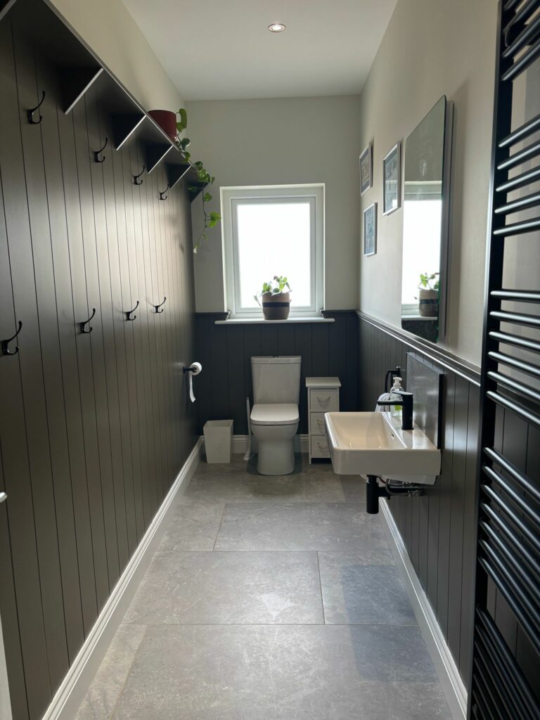 Modern bathroom with a sink, toilet, black paneling, plants on the window sill, and wall hooks.