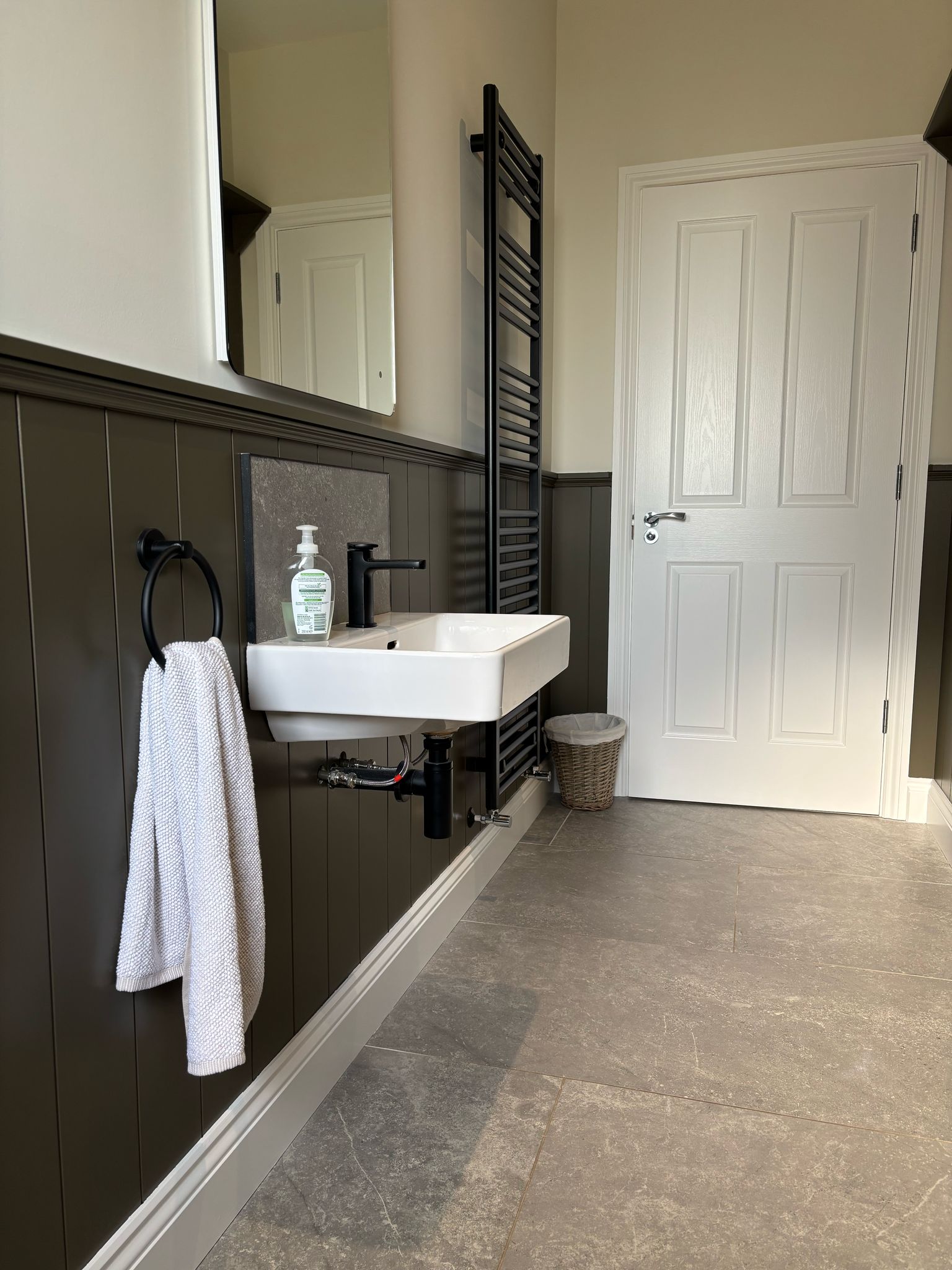Modern bathroom with a white sink, black fixtures, towel, mirror, radiator, and a wicker trash bin on tiled floor.