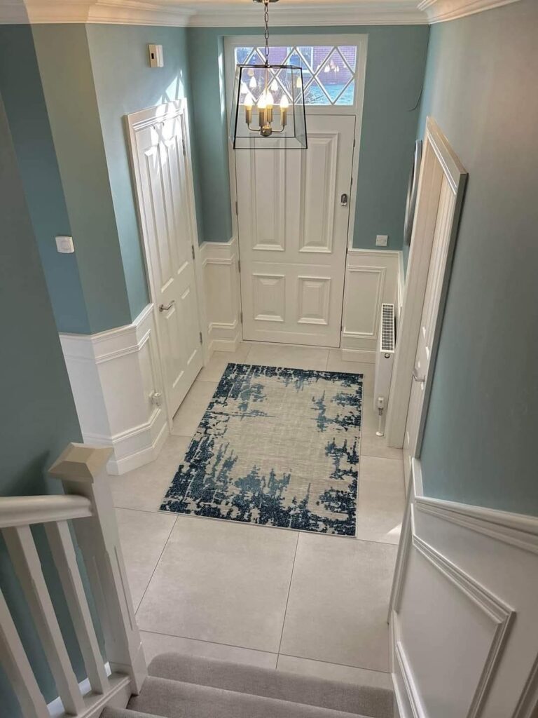 Bright hallway with a white door, chandelier, blue and white rug, and light blue walls with wainscoting.