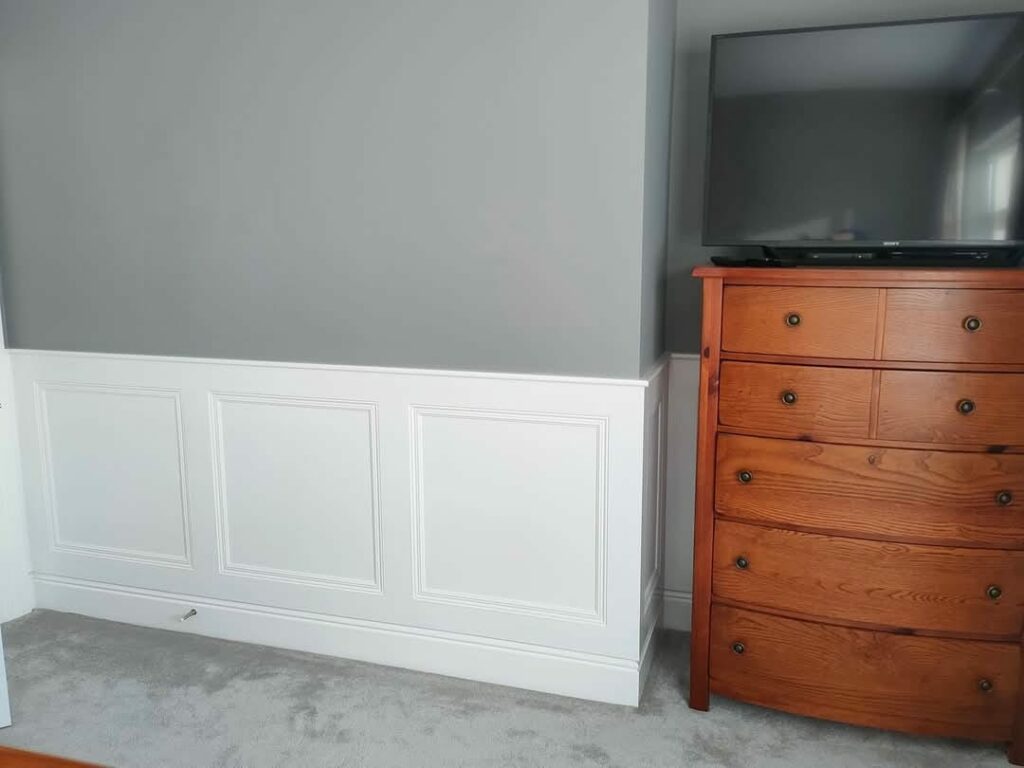 Wooden dresser with TV next to gray and white paneled wall in a carpeted room.