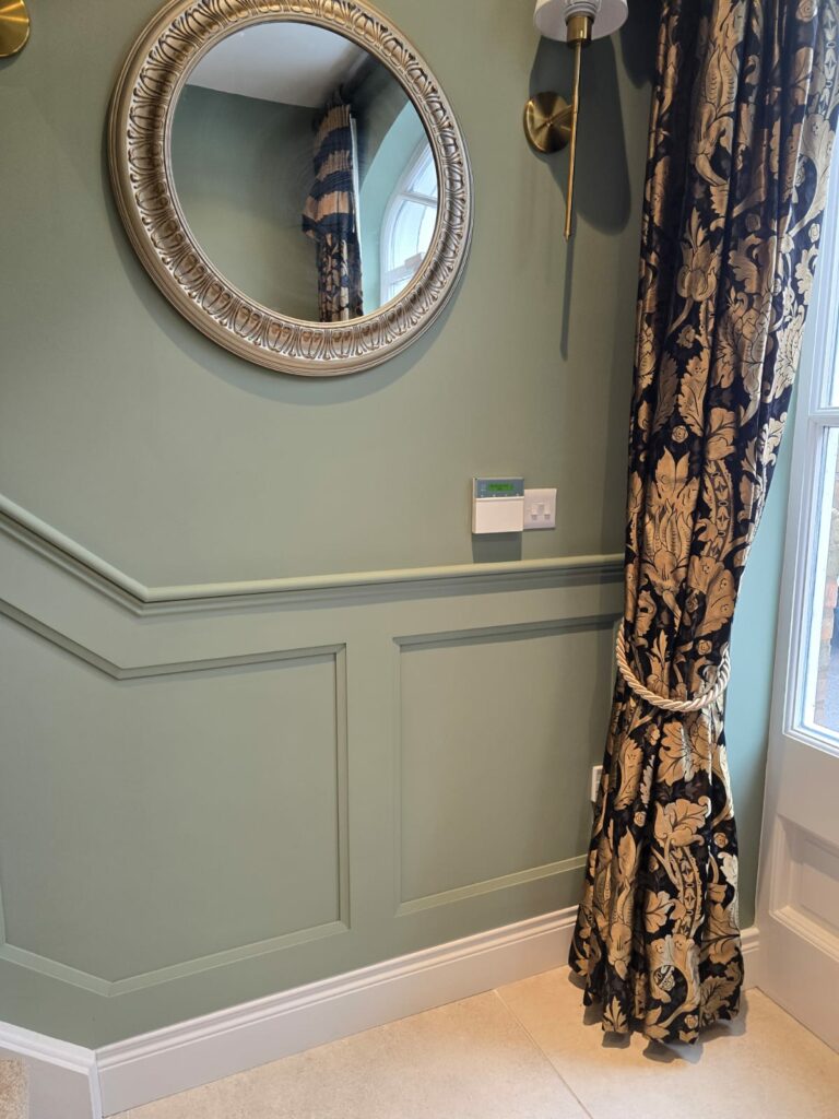 Elegant hallway with ornate mirror, decorative wall paneling, and floral-patterned curtain tied back beside a window.