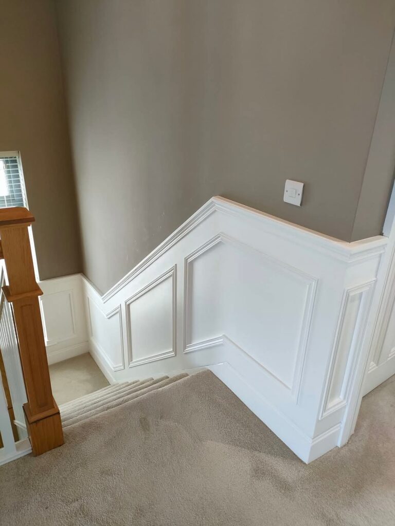Carpeted staircase with white wainscoting and brown walls, featuring a wooden banister and a light switch on the wall.