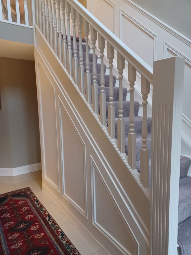 Carpeted staircase with white railings and wall paneling, next to a red patterned rug.