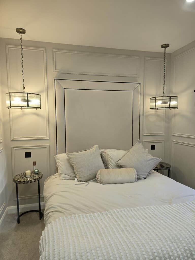 Modern bedroom with neutral tones, featuring a plush bed, two pendant lights, decorative paneling, and side tables.