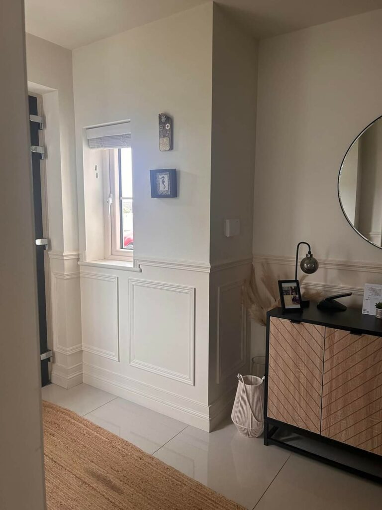 Modern hallway with white wainscoting, decorative wall art, a circular mirror, wooden cabinet, and a woven rug.