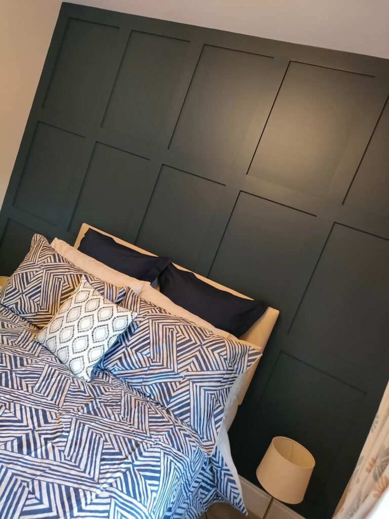 Bedroom with geometric-patterned bedding, dark paneled accent wall, and a small bedside lamp.