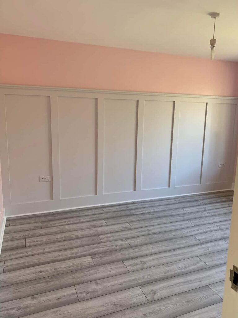 Empty room with light pink walls, white paneling, and gray wood flooring.