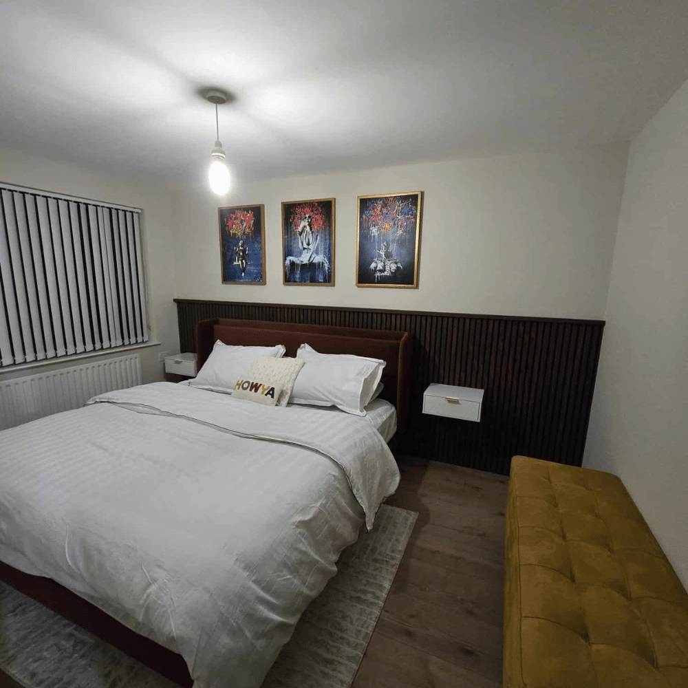 Modern bedroom with a king-size bed, decorative pillows, three framed artworks above, and a yellow bench at the foot.