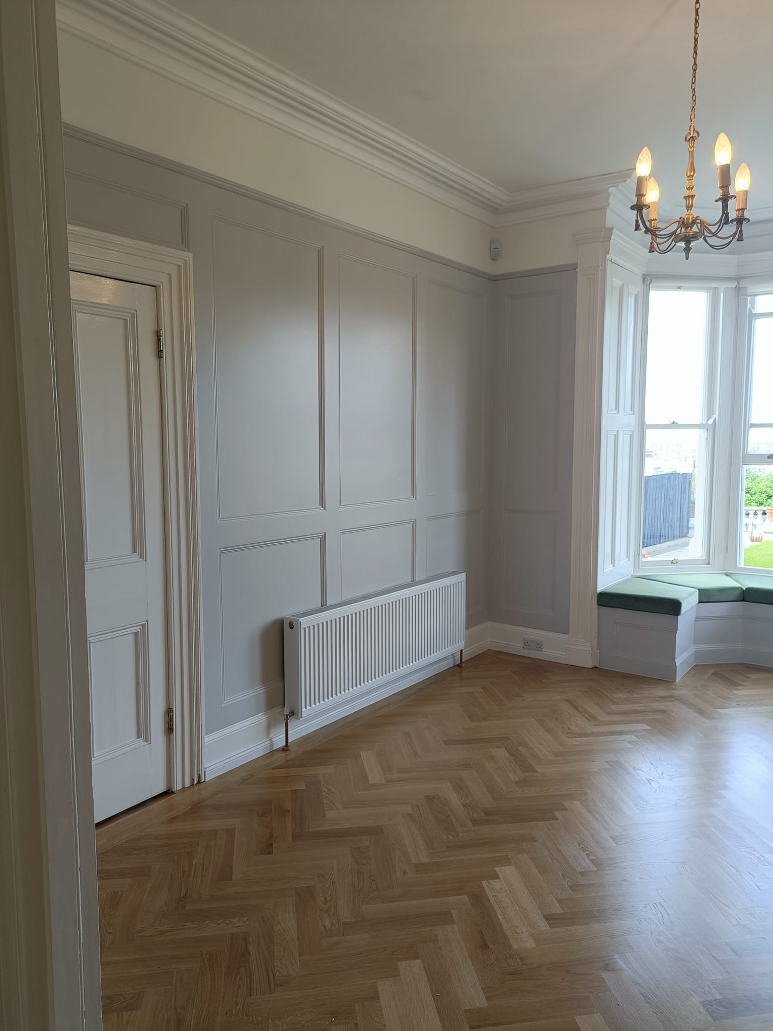 Bright room with herringbone wood flooring, white paneled walls, a radiator, and a window seat under a chandelier.