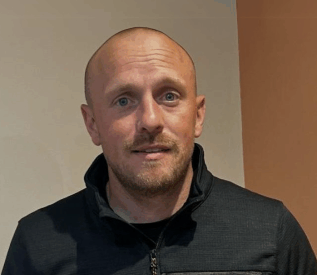 A person with a shaved head and facial hair wearing a zip-up jacket, standing indoors against a two-toned wall.