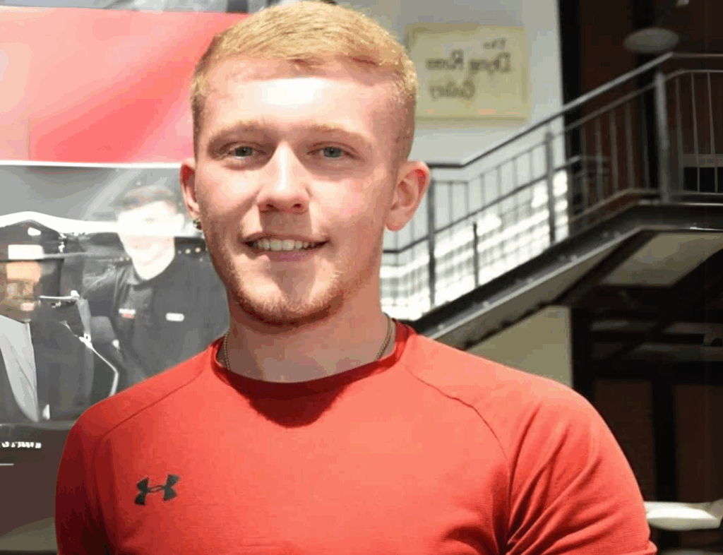 Young man in a red shirt smiling, standing indoors with a staircase in the background.